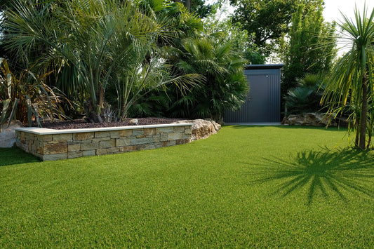 Un jardin aménagé avec un gazon vert parfaitement entretenu, une bordure en pierre surélevée remplie de palmiers luxuriants et de plantes tropicales. En arrière-plan, une cabane de jardin grise avec une porte fermée.