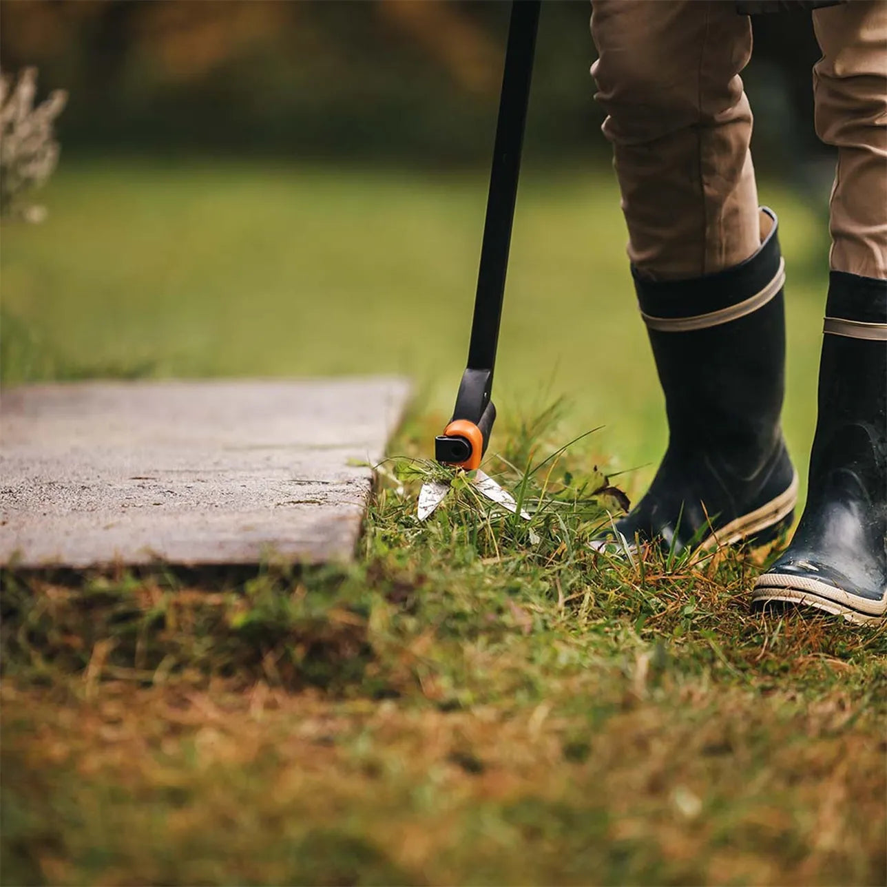 Utilisation des cisailles à gazon longue portée Fiskars pour tailler l'herbe près d'une dalle en béton, avec une vue sur les bottes de l'utilisateur en arrière-plan.