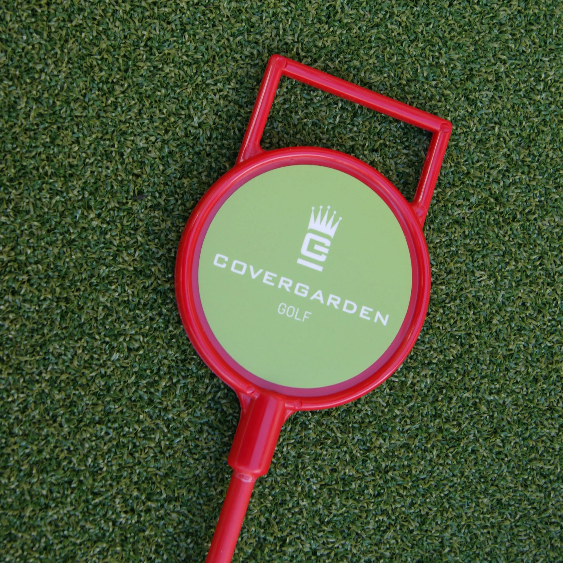 Un drapeau rouge de golf avec une poignée rectangulaire sur fond vert gazon. Le panneau rond affiche le logo 'COVERGARDEN' et le mot 'GOLF'.