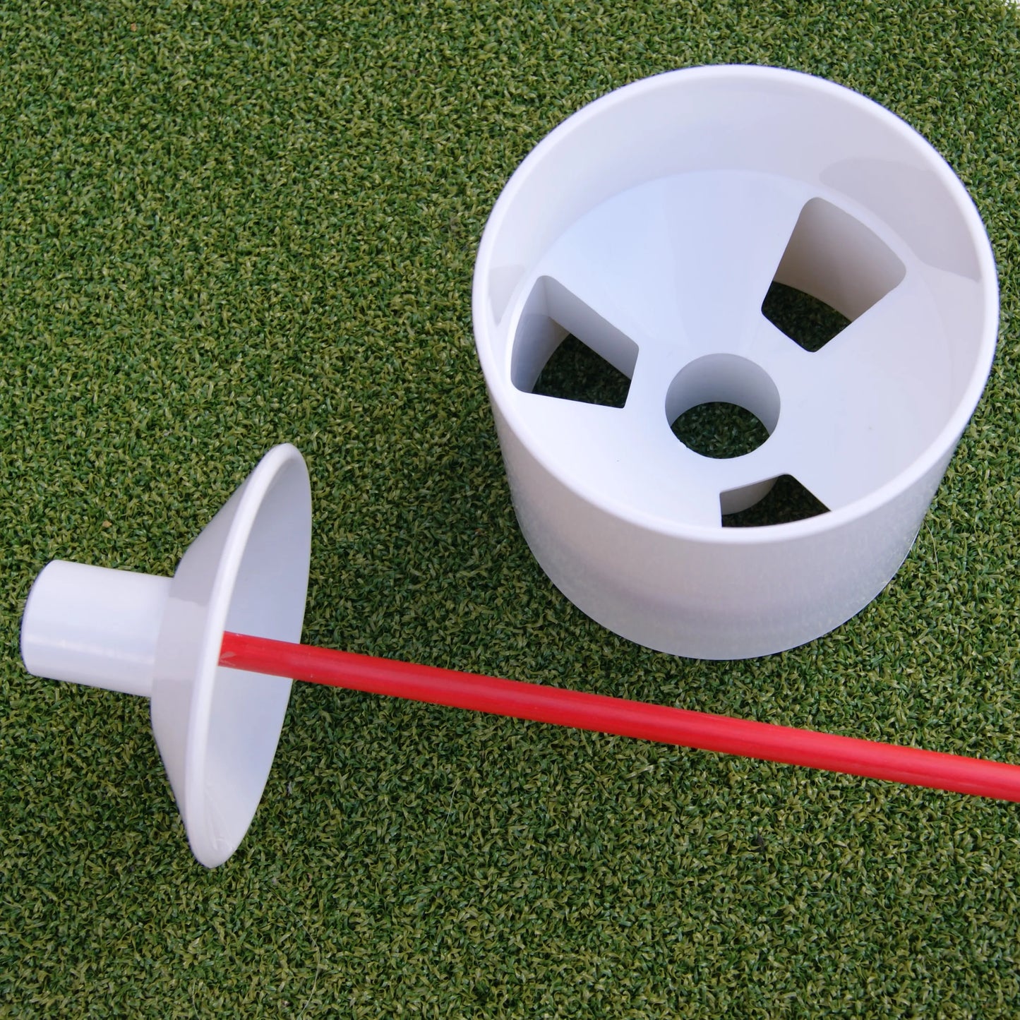 Un gobelet de golf blanc avec des découpes pour laisser passer l'air, placé sur du gazon synthétique, accompagné d'un drapeau rouge en partie visible.