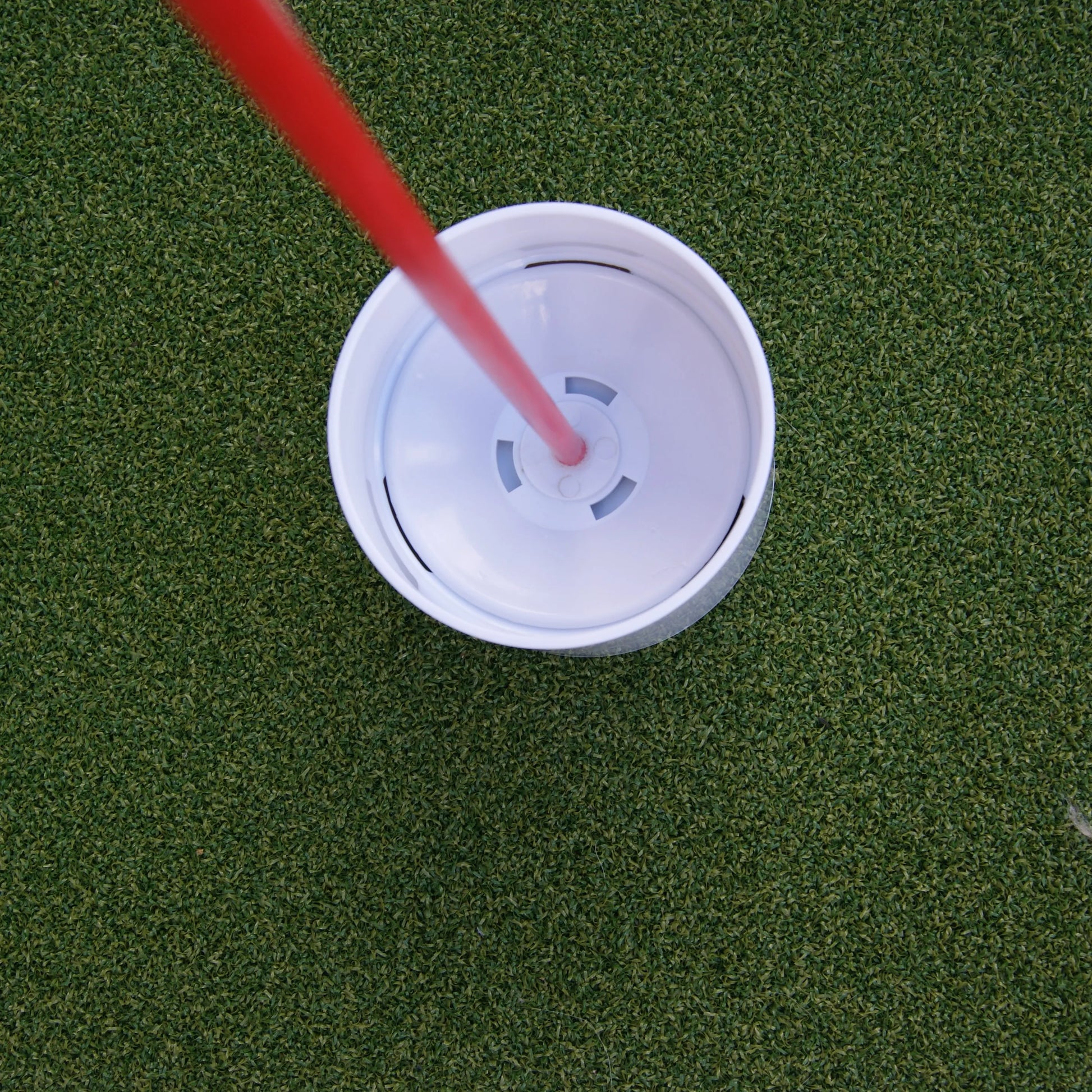 Vue aérienne d'un gobelet de golf blanc avec une tige rouge insérée au centre, placé sur une surface de gazon synthétique bien entretenue.