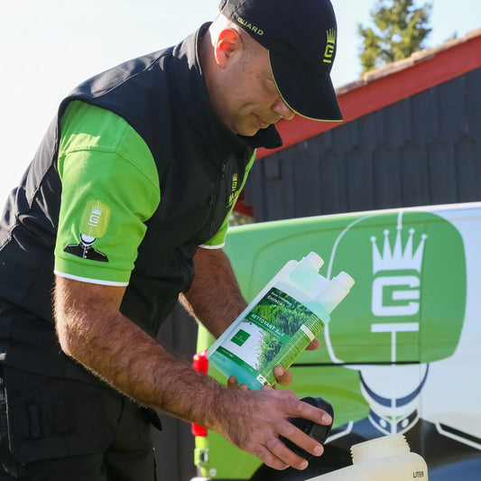 Un professionnel en uniforme COVERGARDEN noir et vert verse un produit nettoyant dans un récipient. Il tient une bouteille de nettoyant gazon synthétique 3-en-1 verte. En arrière-plan, un véhicule de service vert avec un logo blanc COVERGARDEN est visible.