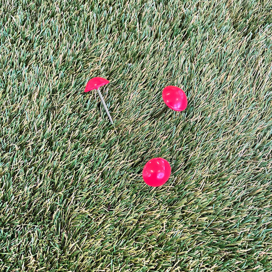 Trois piquets à tête large de couleur rouge vif, disposés sur une surface de gazon synthétique vert. L'un des piquets est planté dans le gazon, tandis que les deux autres sont posés à côté.