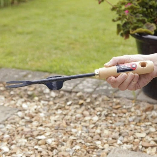 Un outil tire-racine tenu dans une main, avec un manche en bois clair et un embout métallique noir doté d'une fourche à deux dents. L'outil est utilisé sur un sol recouvert de petits cailloux avec un fond d'herbe verte et de plantes en pot.